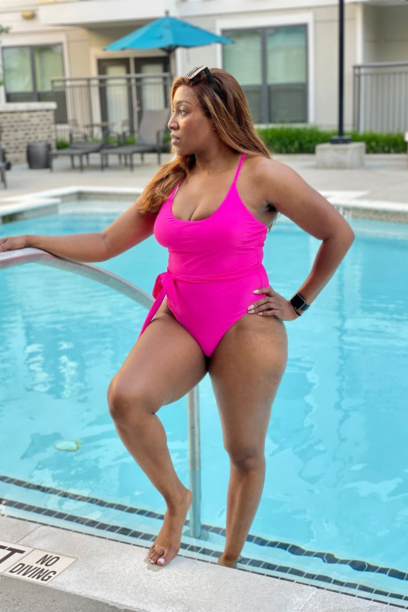 Poolside , woman wearing a hot pink one piece swimsuit with matching belt at the waist. Paired with sunglasses and accented with white toe nail polish.